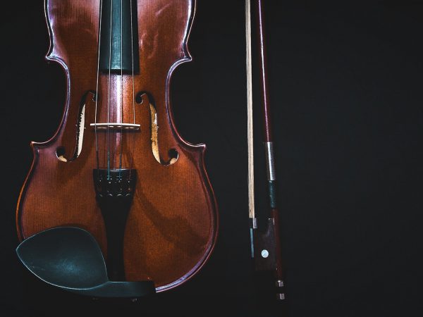 A dark brown wooden violin hanging down with a black backdrop and the violin bow standing next to it.